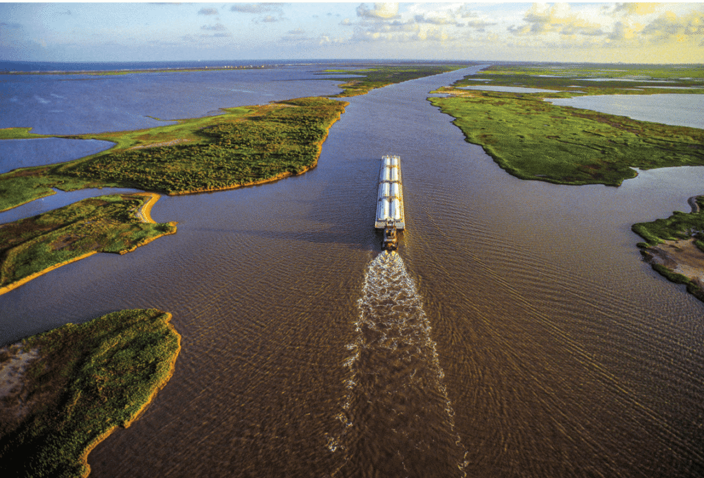 Aerial view of a barge traveling along a wide brown river, surrounded by green islands and water, under a partly cloudy sky (Gulf Intracoastal Waterway expansion).